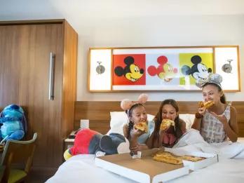 Three young girls sat on a bed eating pizza from a takeaway pizza box