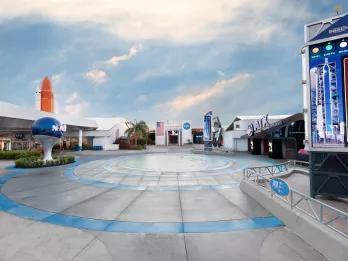 A paved plaza surrounded by space themed buildings, with a large model of the NASA logo to the left-hand side