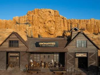 A wooden barn-like building in front of sandy cliffs with a carved Aztec face in the centre