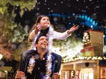 A girl sitting on a man's shoulders holding her hands out to catch fake snow as it falls