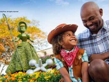 A young boy dressed as a cowboy smiling up at his dad. They are both sat in front of a topiary shaped like Bo Peep from Toy Story