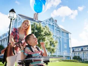 A woman pushing a smiling boy in a wheelchair in front of a blue and white hotel building. There is a blue Mickey-shaped balloon attached to the wheelchair