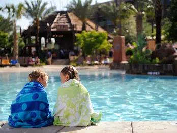 Two young children sat at the edge of a pool wrapped in colourful towels