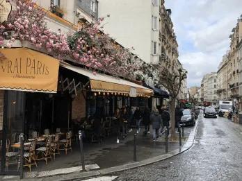 A wet road in Paris lined with parked cars and the outside of s restaurant which is named "Vrai Paris". Above the beige cover there are pink flowers and below there are tables with chairs.