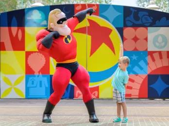 A young boy posing with Mr Incredible in front of a gate painted with the Pixar Ball