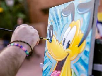 Man painting on canvas and the painting is of Pluto the dog, a yellow cartoon dog, with a blue background.