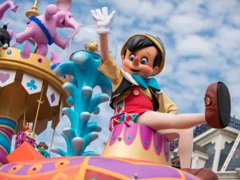 Cartoon puppet made from wood waving from above a float. Pinocchio is sat on a seat at the front of a large float full of statue elephants in tutus on top of a mini statue of kids sat on a swing ride.