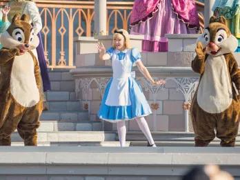 Alice from Alice in Wonderland stood on stage waving at guests, wearing her iconic blue dress with a white apron over, black bow in her hair, and white tights. Next to her either side are the two cartoon chipmunks, one with a black nose, the other with a red nose.