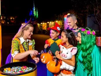 A woman wearing a bat headband and a cast member nametag handing out sweets to guests who are dressed as Hei Hei, Moana and other Moana characters. The young boy is wearing a cap resembling a chicken and the girl is wearing a moana costumer and holding a plush of Pua the pig.