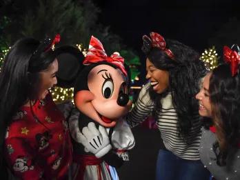 Minnie mouse stood in a white and red stripey bow and a matching dress. There are three women crowded around her, one on the left wearing a red jumper with festive Disney cartoon characters on and the other two on the right wearing jumpers and jeans and sparkly Minnie mouse ear headbands.