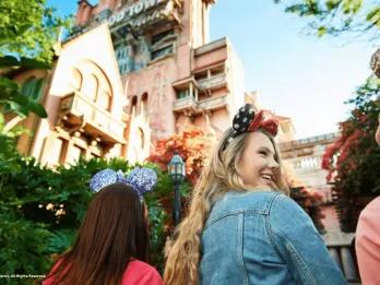 A group of young girls stood wearing mickey ears underneath a large dishevelled looking tower which is the Tower of Terror. One of the girls is turned to the others smiling. 