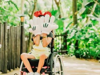Young child in a wheelchair covering their face with two hands which have large white mickey gloves on. They are wearing red sparkly mickey ears on their head. In the background there is a pathway with trees surrounding it.