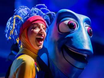 A woman with a yellow full body suit which turns red on her head. On the top of her head she has clue strands coming off which looks like pieces of a coral reef. To the right is a large puppet of Dory, a blue fish staring in the same direction as her. 
