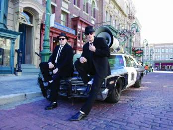 Two men in black suits and hats are leaning against a black and white car which has a large speaker on top of it. They are in the middle of the New York set streets.