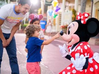 Large cartoon mouse wearing a red dress with white polka dots on it and a matching bow. There is a young boy touching Minnie Mouse's nose. There is a young girl next to the boy and a man bending down smiling and watching. In the background there are multi-coloured Mickey balloons.