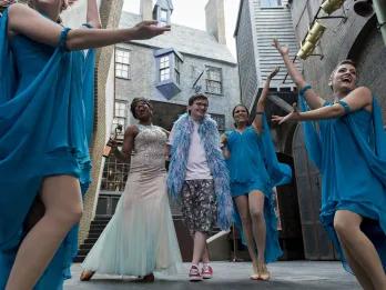 Woman in a pretty, elegant floor length white and blue dress singing with a man next to her wearing a blue feather boa. Around there are three women wearing shorter blue dresses.