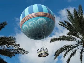 Large blue hot air balloon flying up in the sky with two palm trees either side.
