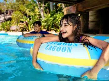 Little girl and little boy smiling inside of rubber rings on a lazy river. They are floating under a bridge and there are many trees behind them.