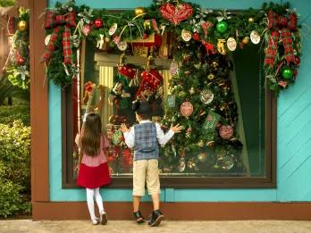 Two kids, one boy and one girl, stood in shop window looking inside wearing Mickey Mouse ear shaped hats. Inside there is a Christmas tree lavishly decorated and above the window there are christmas decorations.