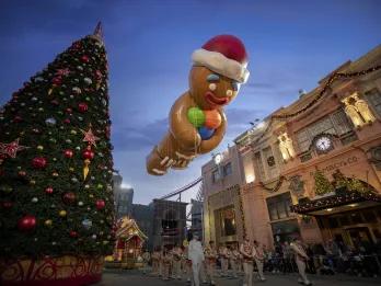 Huge Christmas tree with pretty decorations on. Next to it is a giant gingerbread man in christmas Santa Claus hat floating in the air.