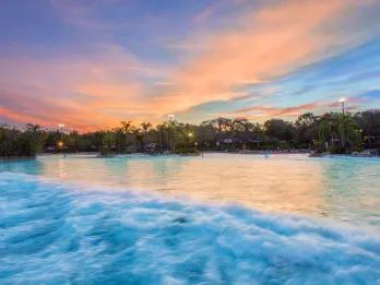 Sun set with a blue sky and pink/orange coloured clouds. There is a huge pool with a wave splashing through which is surrounded by trees.