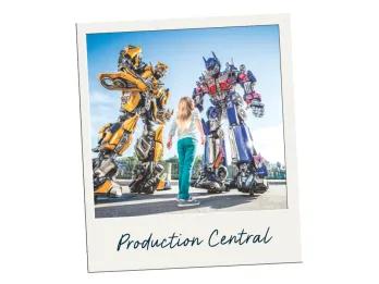 Young girl stood looking up to two giant robots, one yellow and the other blue and grey. They are Transformers. The polaroid is labelled Production Central.