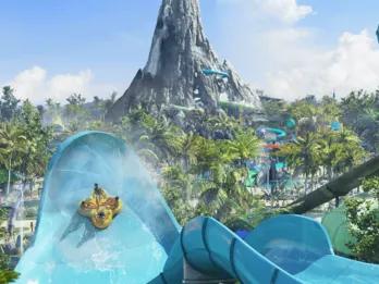 Four people facing each other on a large yellow rubber ring flying up a light blue water slide wall which is huge. In the background, there is a volcano statue with slides coming off of it and water.