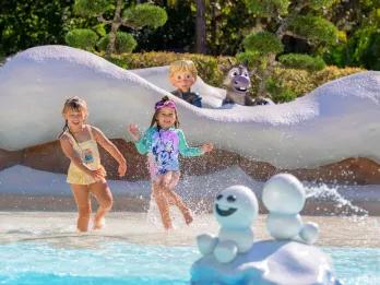 Two little girls playing in a shallow pool. Behind there are snow walls and a statue of young Kristoff and Sven are poking over from Frozen. There are also statues of the small snow ball shaped snowmen in the middle splashing water.