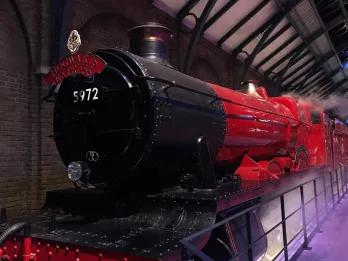 Large steam train with the label Hogwarts Express, coloured black and a dark red. There is steam surrounding and it is parked in a large factory type building.