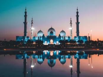 A beautiful religious temple in Abu Dhabi located overlooking a lake. The sun is setting with a pink and blue hue in the sky and the temple is perfectly reflected in the lake. It is a white building with three white domes and four big pillars sticking out.