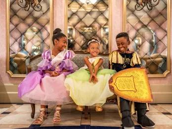 Three young children sitting on a couch dressed as princesses and a prince
