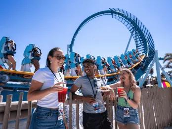 Three young adults stood in front of the Pipeline rollercoaster drinking cocktails and eating festival plates