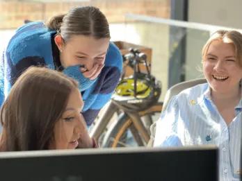 Three girls gathered around a computer laughing, with one covering her mouth with her jumper