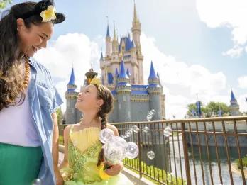 Mum holding the hand of her daughter who is dressed as Princess Tiana and holding a Mickey shaped bubble wand. In the background there's Cinderella Castle.
