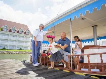 Family getting off of a small boat wearing mickey ears and holding merchandise. In the background there is a white building hotel.