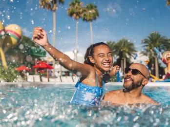 Dad lifting daughter up in the resort pool, both smiling. There are giant statues of musical instruments.