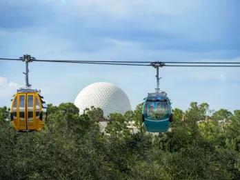Two cable cars, one orange and one blue, moving along cables in front of Spaceship Earth, a giant golf ball looking building.