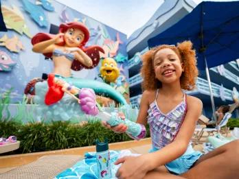 Girl sat on a sunbed holding an Ariel bubble wand in front of a giant statue of Ariel sat with flounder on the side of the hotel.