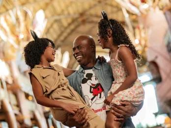Man wearing a Mickey Mouse shirt holding two girls who are wearing safari style outfits and Mickey Mouse ears.