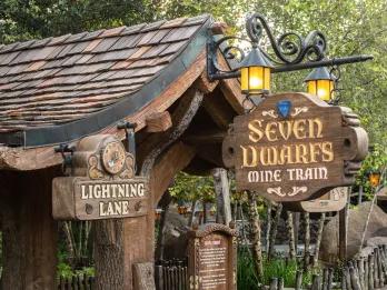 Small wishing well themed entrance to a ride with brick roof and a wooden sign saying "seven dwarfs mine train". To the left there is a sign saying Lightning Lane. 