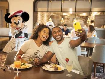 Man taking a selfie with his girlfriend smiling at a restaurant. In the background Minnie Mouse is posing with an artist outfit on.
