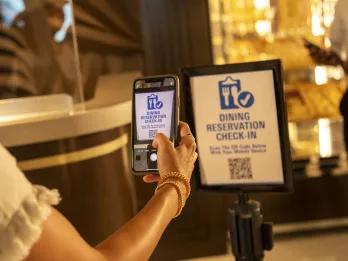 Poster with checkboard and a knife and fork cartoon icon and check mark. It says dining reservation check in and someone is holding their phone up to scan a QR code.