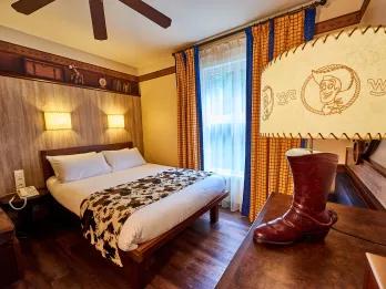 Standard room with wooden floor, yellow curtains and a double bed. It is themed like the wild west with cow print blankets over the bed and a lamp with woody from toy story on it and a cowboy boot.