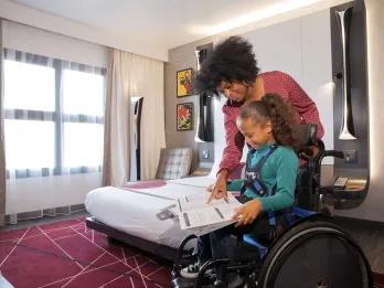 Hotel room with burgundy carpet and double bed and a girl in a wheelchair reading a book and her mum stood over her.