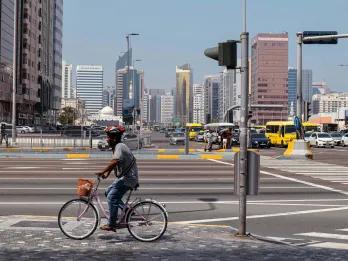 Man cycling on the road with many cars and buses in the background. There are also many modern high rise buildings lining the street.