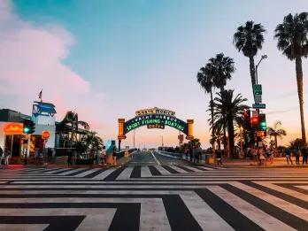 Zig zag black and white lines on the floor leading up to a large sign for Santa Monica pier lighting up as the sun sets. There are palm trees next to it.