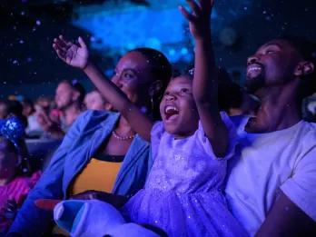 A little girl smiling and holding her arms up while sat with her mum and dad in a theatre