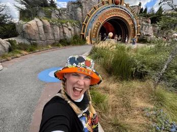 Woman in a bucket hat which has text on it saying Monsters and cartoon art on it. She's stood with her mouth open in excitement in front of one of the Epic Universe portals.