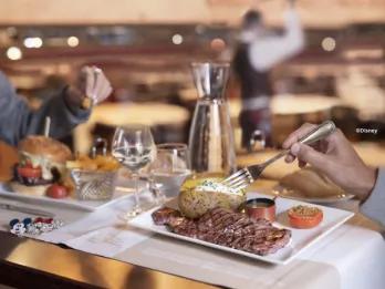 A grilled steak served with a baked potato and dipping sauce on a white plate at The Steakhouse in Disney Village, with wine glasses and a burger in the background in a warm restaurant setting
