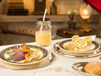 An elegantly presented American meal at Walt's restaurant in Disneyland Park, featuring a main plate of grilled fish with vegetables, a Mickey-shaped waffle dessert, a creamy milkshake in a mason jar, served on vintage-style silver crockery
